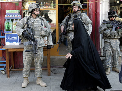Three soldiers in camouflage uniforms and gear stand on a sidewalk in front of a store. A person in a black robe, likely a woman, is walking past them. The store has colorful products visible through the window.